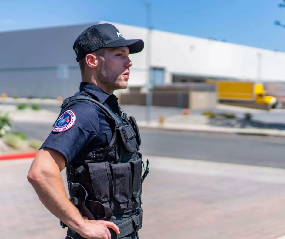 A male security officer in uniform standing outdoors under sunny weather with a serious expression, wearing a black cap and tactical vest, with a security patch on his sleeve.
