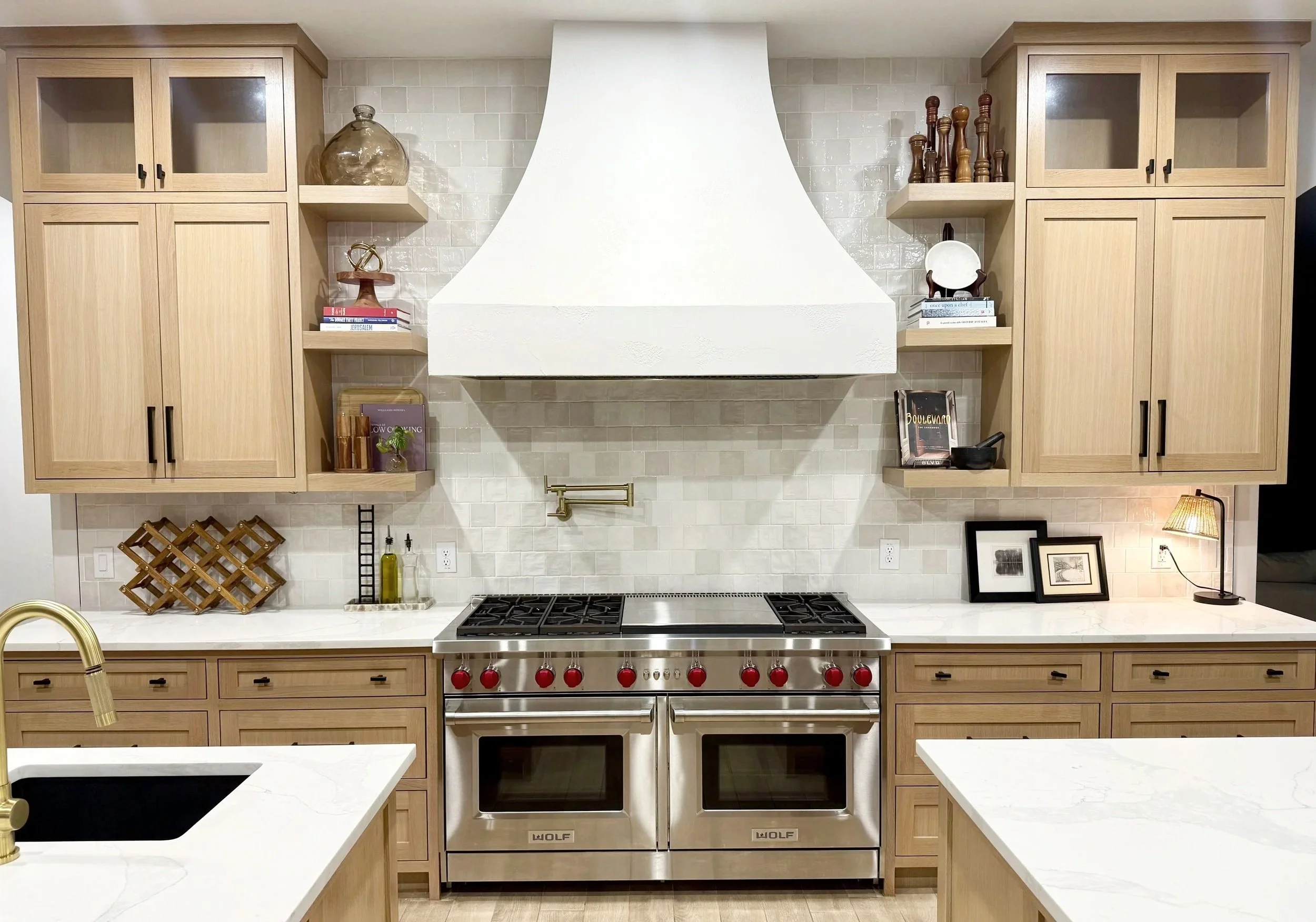 Modern kitchen with wooden cabinets, a large stainless steel stove with red knobs, white countertops, and a white range hood. Decor includes books, framed photos, and small decorative items.