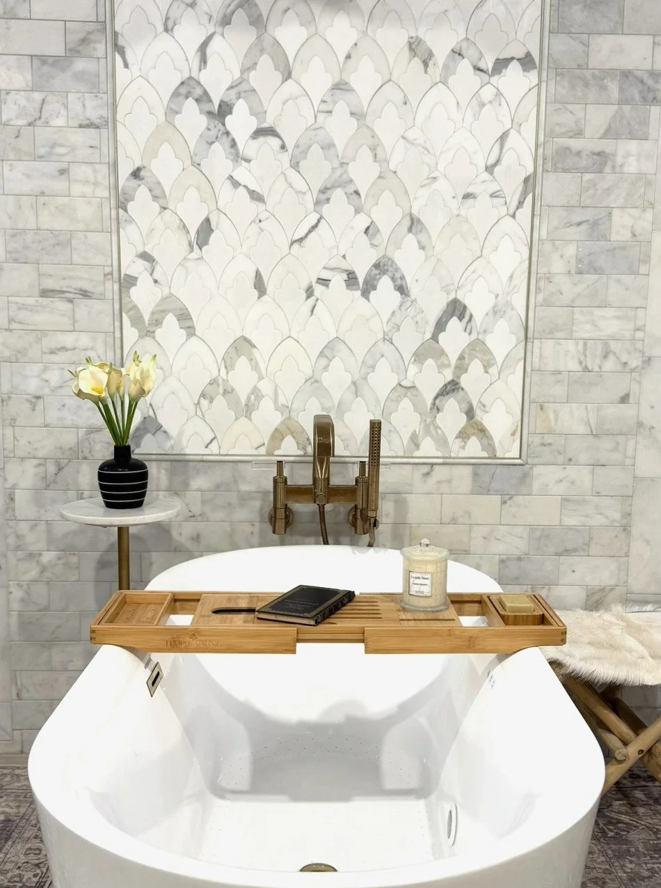 Modern bathroom with a round bathtub, a wooden bath tray with a book and candle, a black vase with white calla lilies on a small white table, decorative tile wall, and a fur-covered stool.