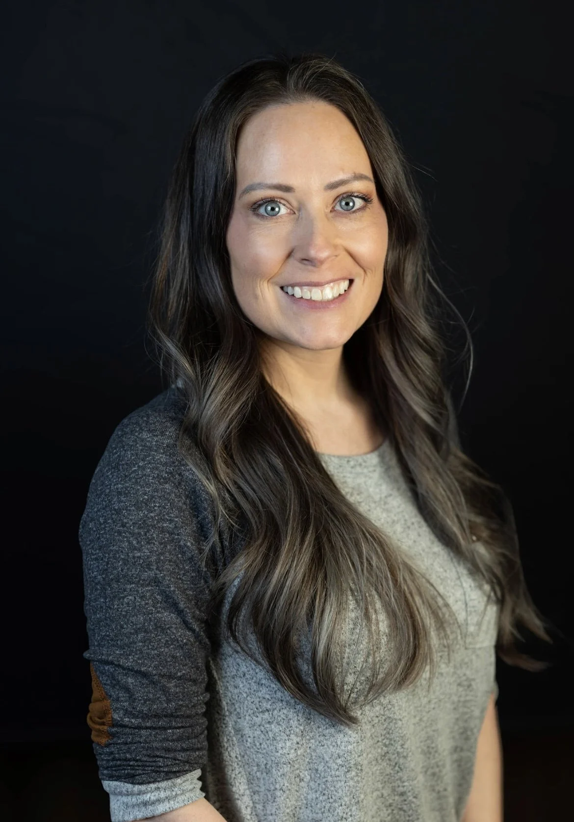 A woman with long wavy brown hair, blue eyes, and light skin, smiling, wearing a gray shirt with darker sleeves, standing against a dark background.