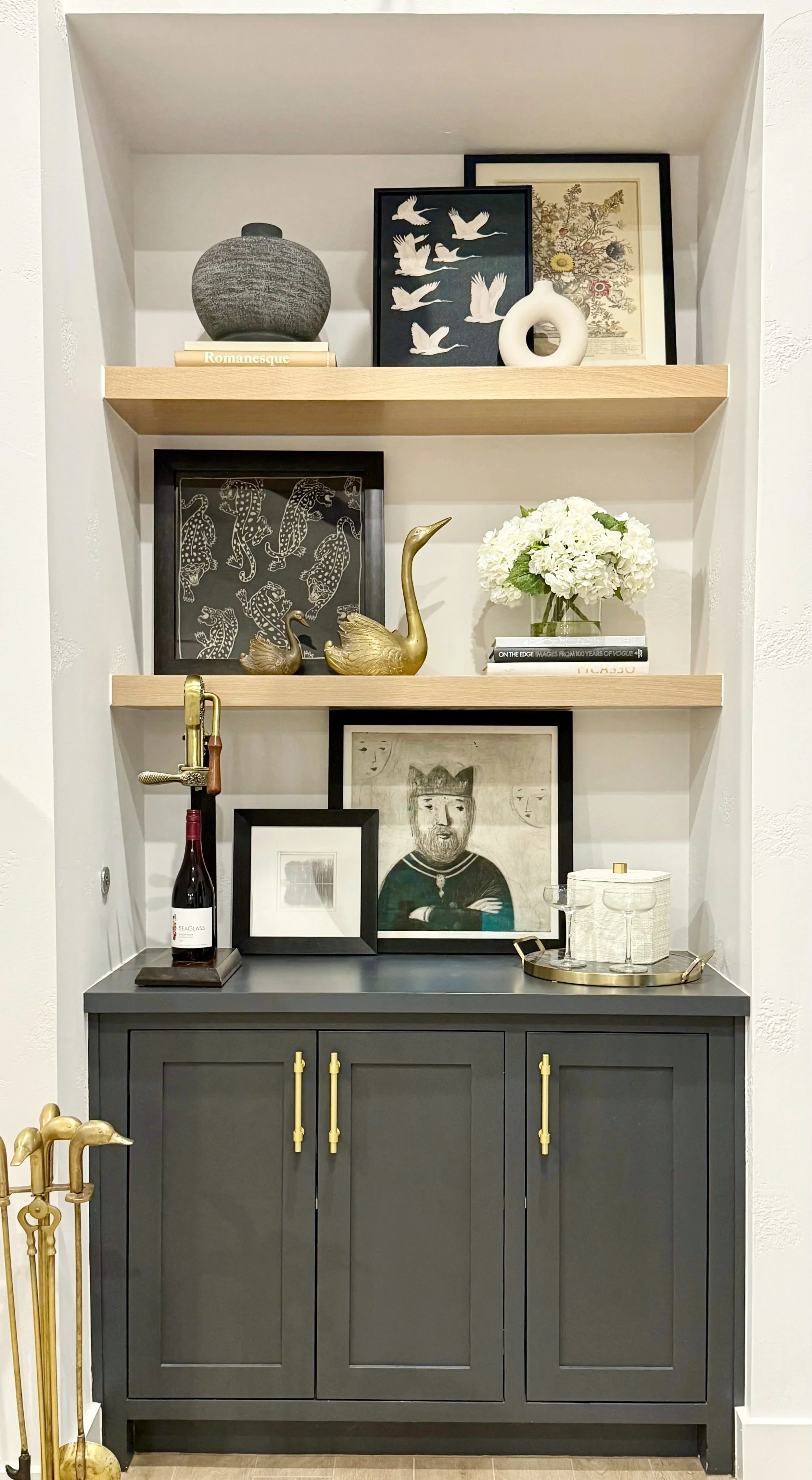 Decorative built-in wall shelf with three wooden shelves displaying art prints, vases, and decorative objects, with a dark gray cabinet at the bottom holding framed pictures and a small tray with glasses and a bottle.