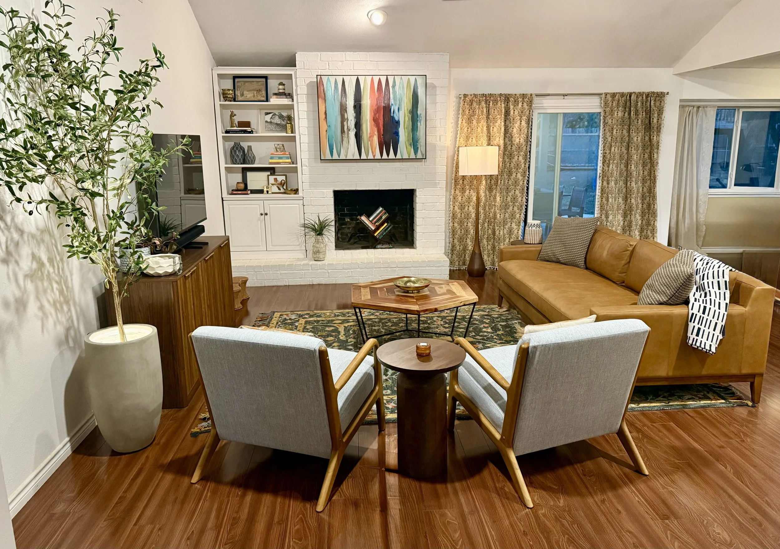 Living room with a white brick fireplace, brown leather sofa, light blue armchairs, a wooden coffee table, and a decorative rug. there are curtains and a large window behind the sofa, a potted plant, and a TV on a wooden cabinet.