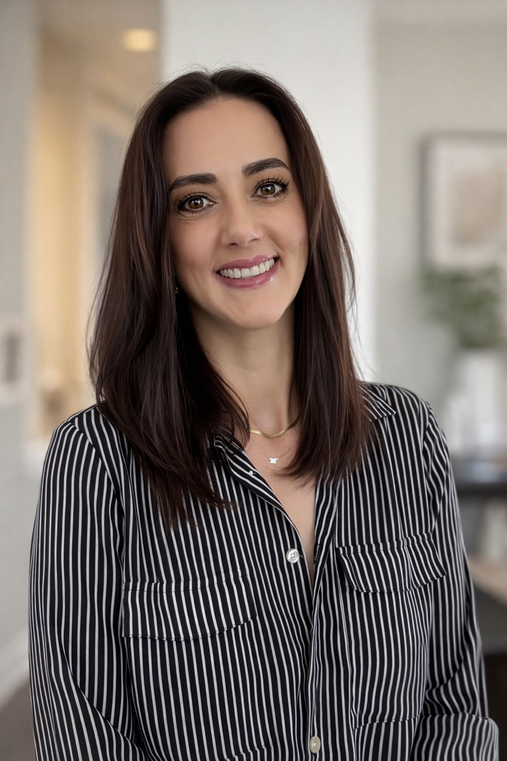 A woman with long brown hair, wearing a black and white striped shirt, smiling in an indoor setting.