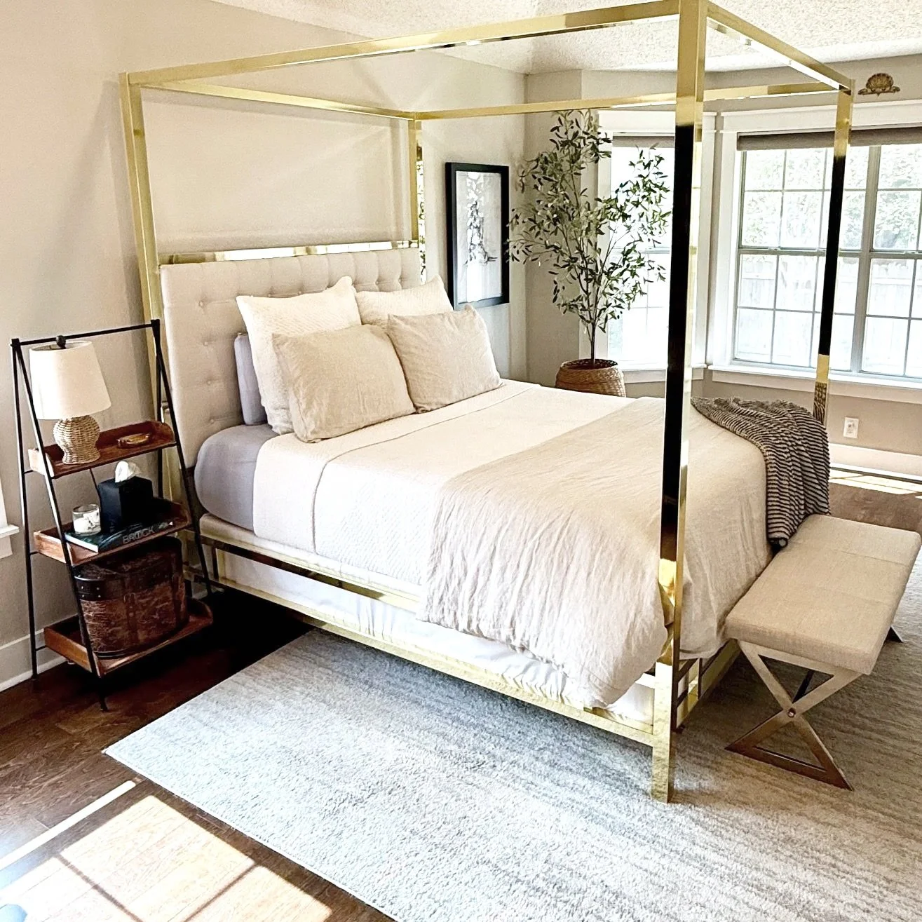 Bedroom with a four-poster bed with a tufted headboard, beige bedding, and pillows. A small bench at the foot of the bed. A side table with a lamp, books, and decorative items. A potted plant near the window, which has two large panes and white trim. A framed picture on the wall.