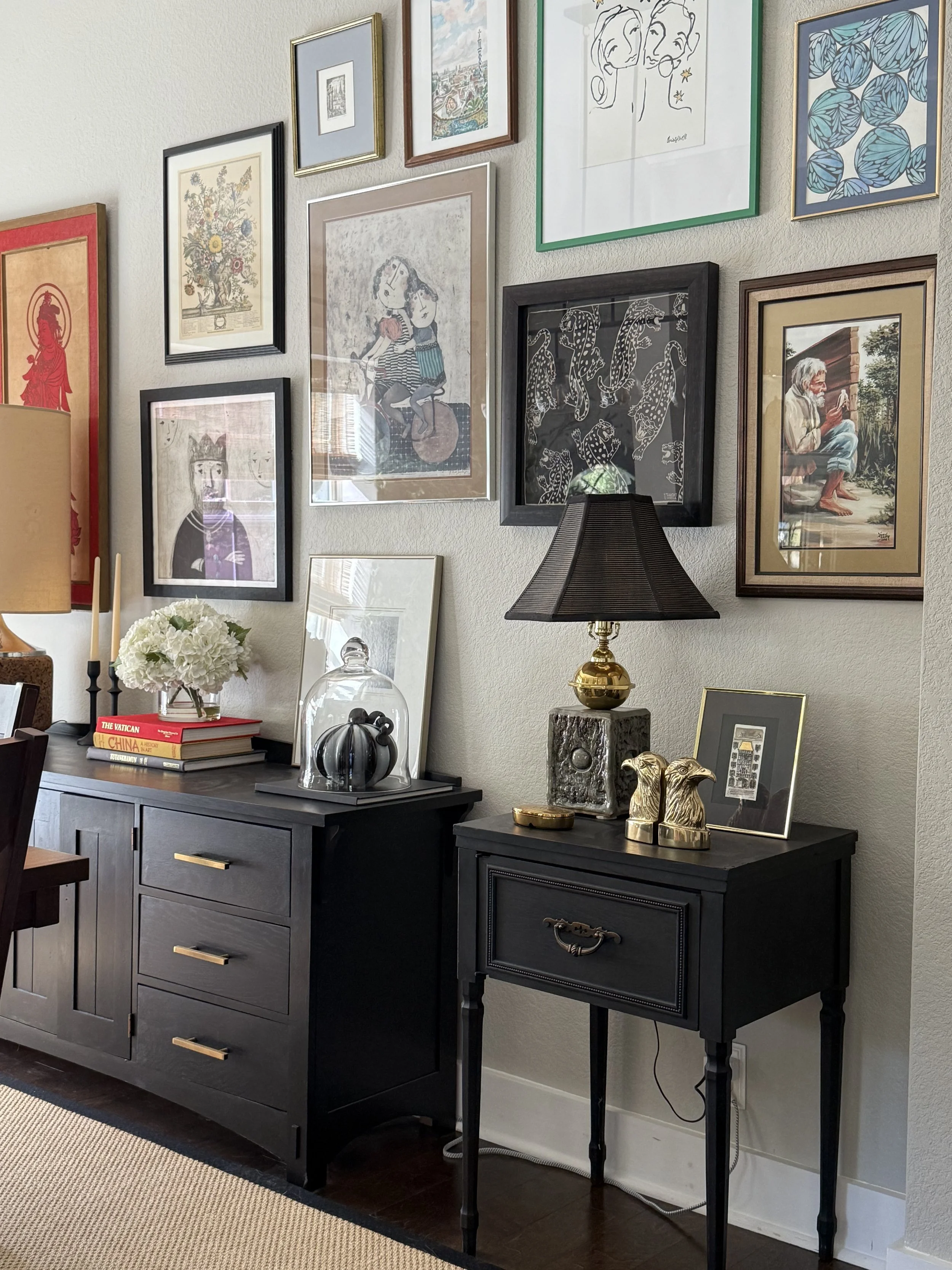 Living room corner with wall art gallery, black sideboard, and black side table. Wall art includes various framed prints and paintings. Decor on side tables includes books, a flower arrangement, a glass cloche with pumpkins, a table lamp, and decorative brass animal figures.