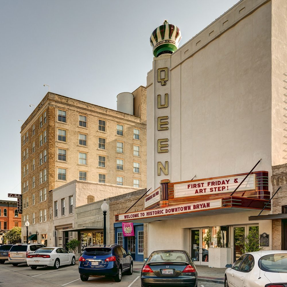 Downtown Bryan, Texas, with a theater marquee reading 'First Friday & Art Step! Welcome to Historic Downtown Bryan.' Several parked cars line the street near historic buildings, and a tall sign spells out 'QUEEN' on the theater building. The sky is clear, indicating a sunny day.