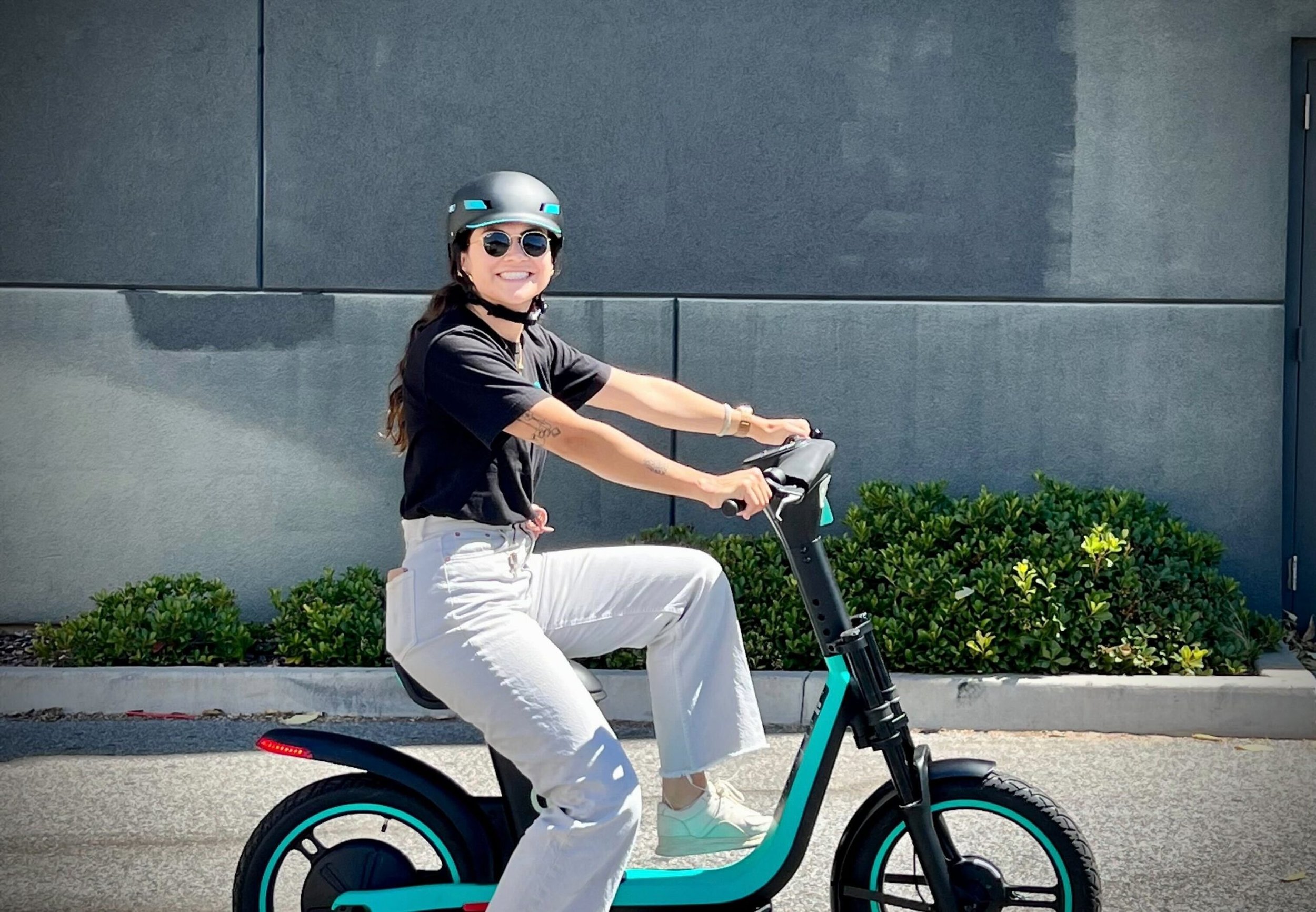A woman wearing sunglasses, a black helmet, a black t-shirt, and white pants rides an electric bike on a city street, smiling at the camera.