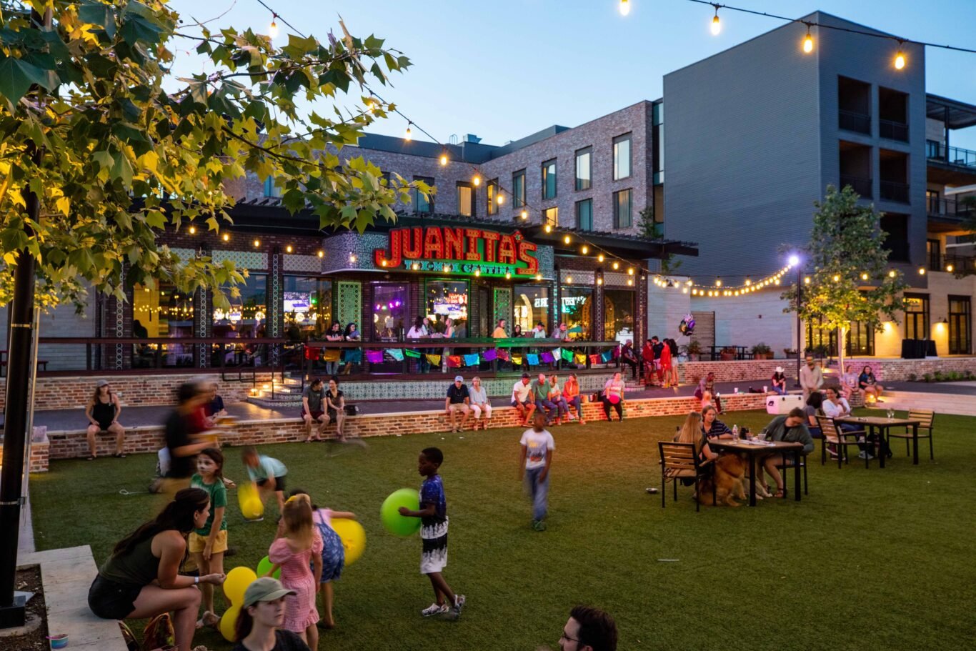 An outdoor scene of Juante's Tex-Mex restaurant with people dining and children playing on a grassy area at dusk, string lights overhead, in an urban setting with modern buildings.