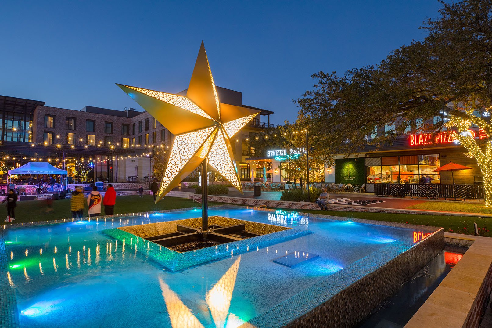 A large illuminated star sculpture is placed in the center of a fountain at an outdoor shopping or dining area during evening. The surrounding area features lights, trees, and various storefronts including a sweet shop and a pizza restaurant.