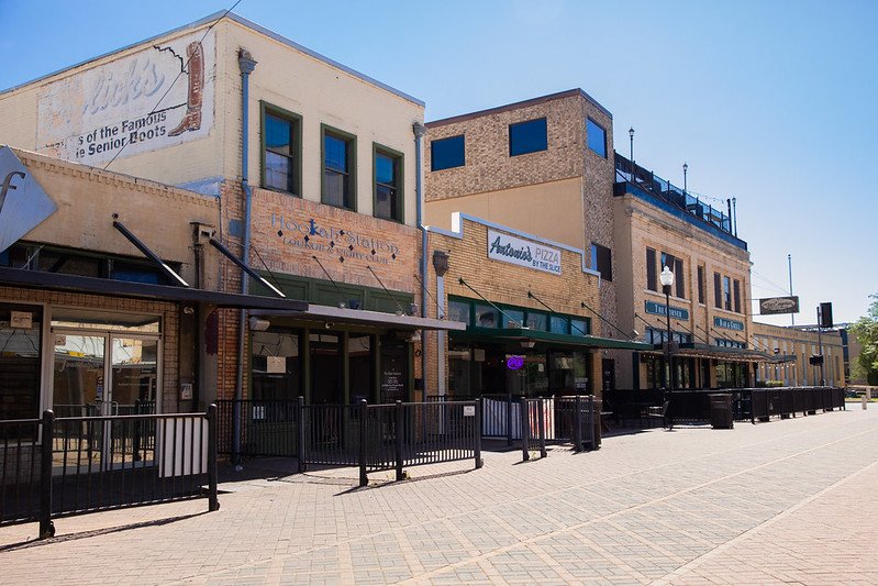 Empty storefronts and restaurants along a pedestrian street in a small town, with clear blue skies and no people visible.