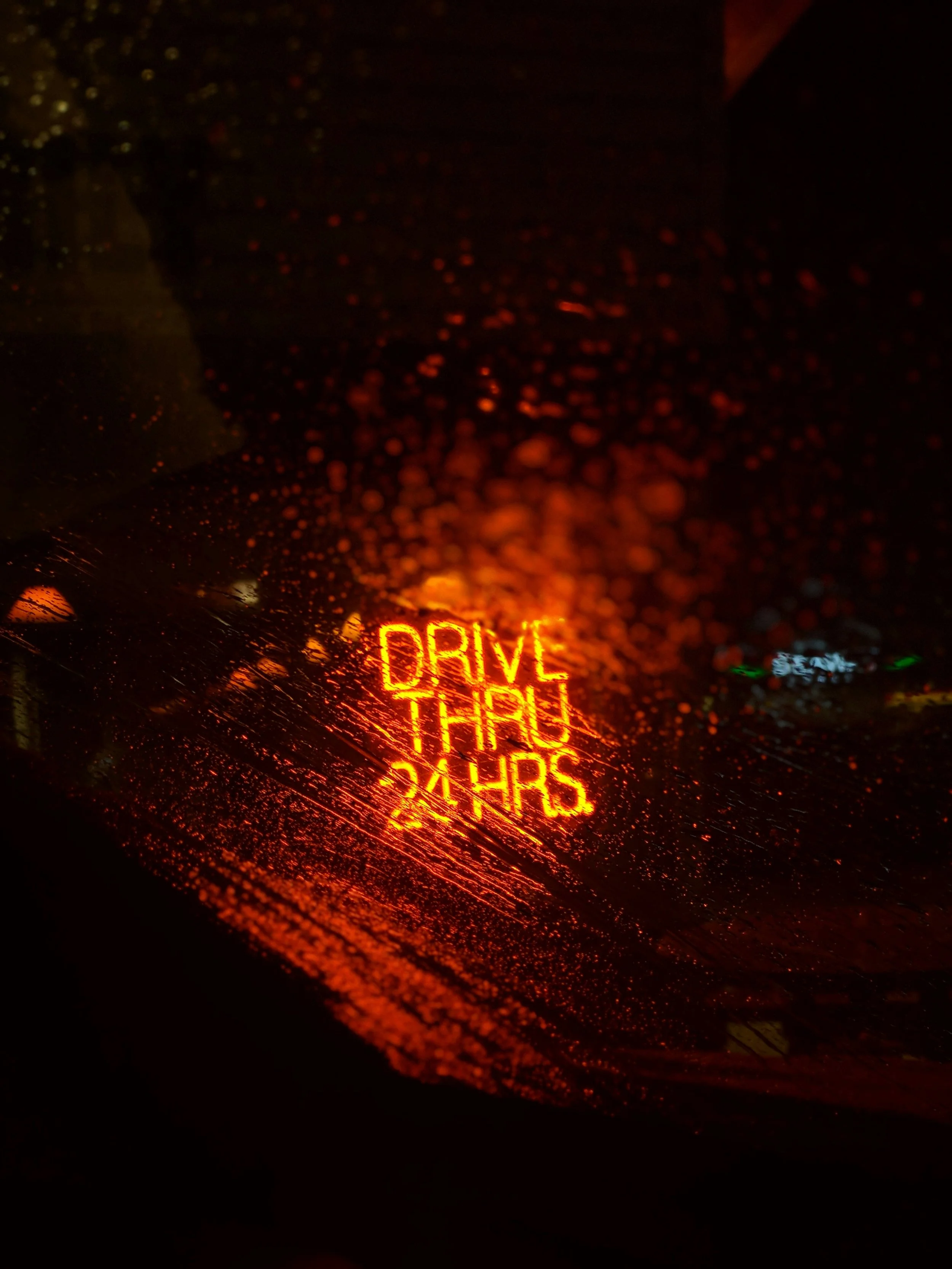 Rain-covered car window reflecting a neon sign that reads 'DRIVE THROUGH 24 HRS' in bright red and orange colors.