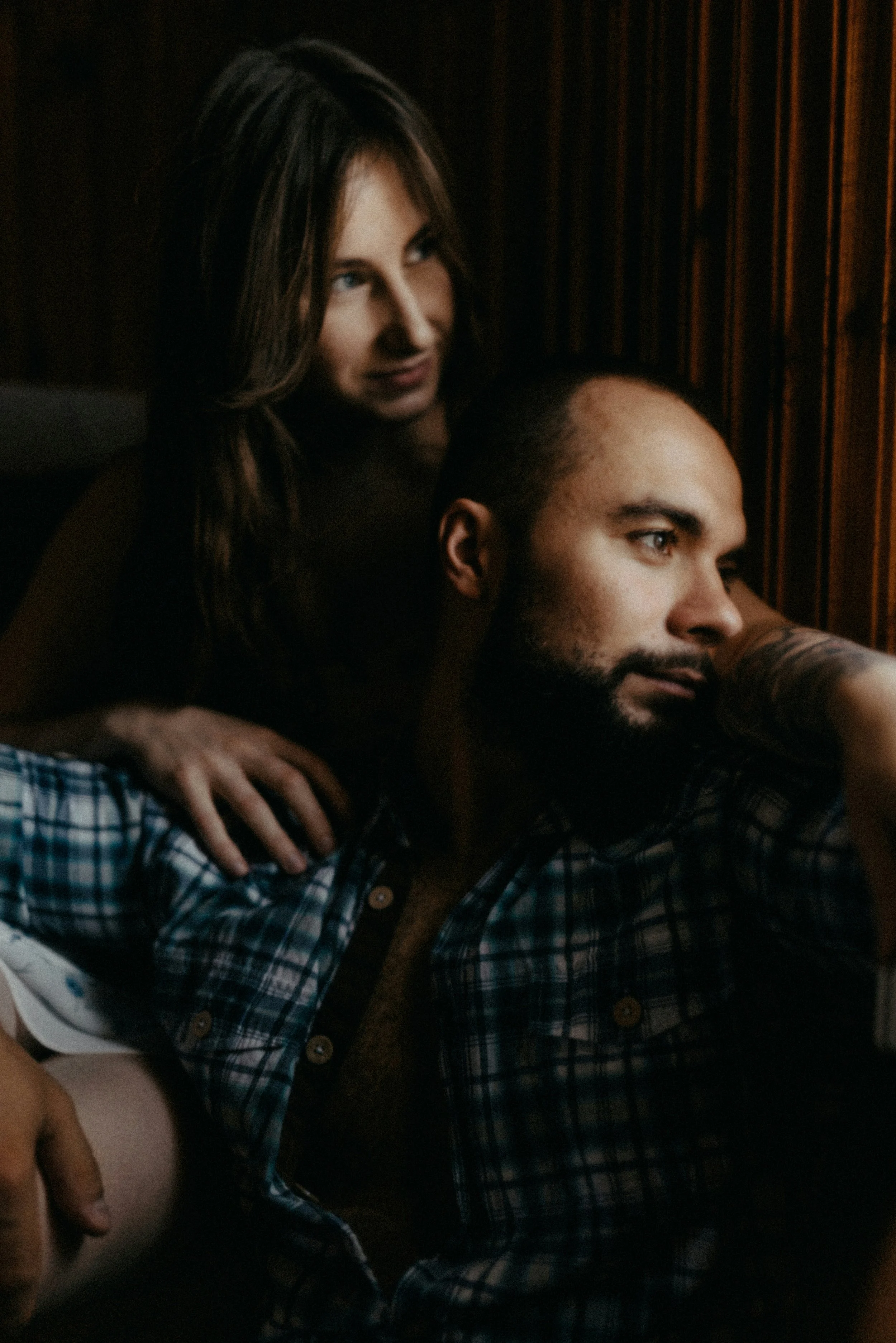 A woman with long brown hair leaning over a seated man with a beard and short hair, looking at something off-camera. She is resting her hand on his shoulder in a dimly lit room with wooden paneling.