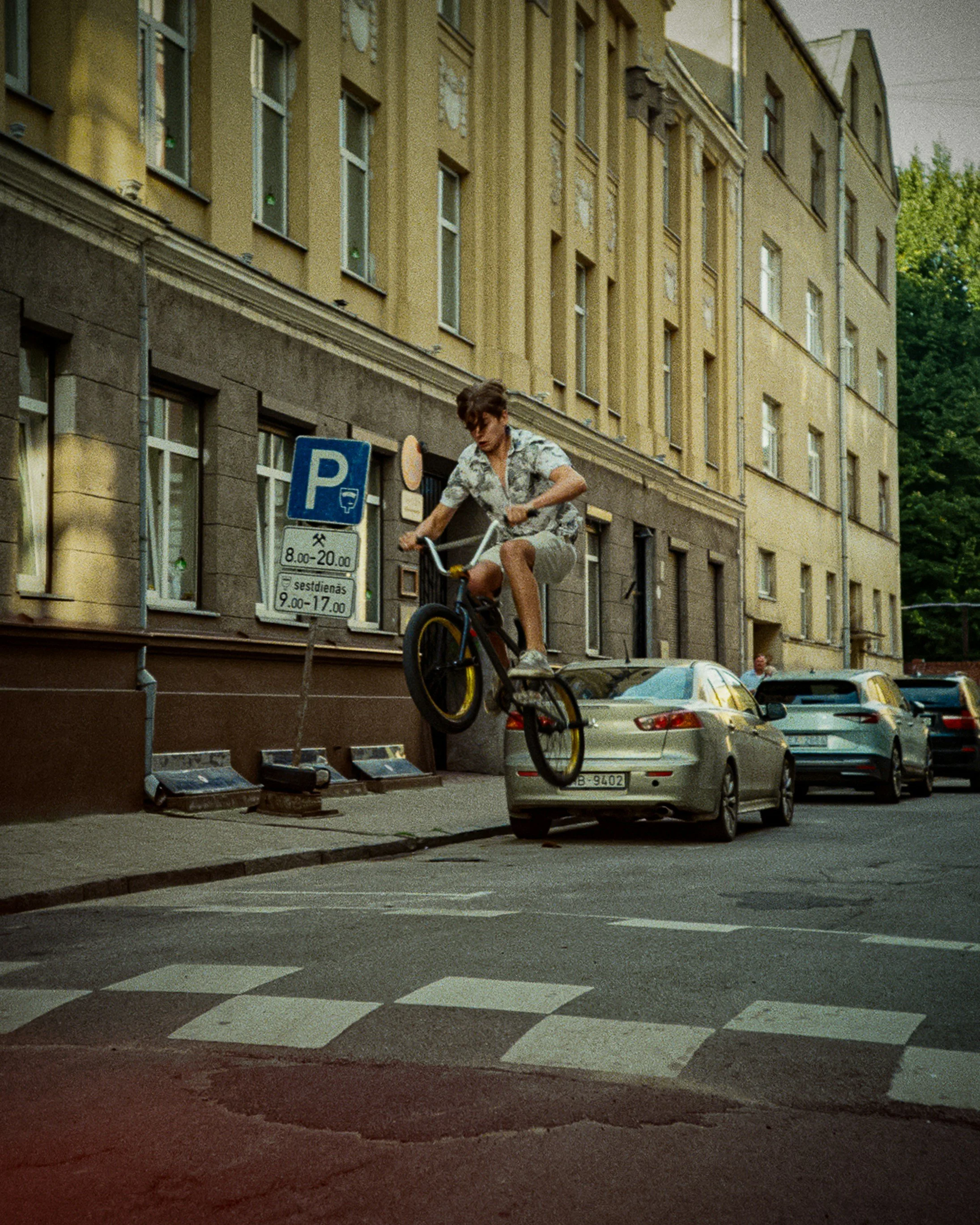 A person performing a BMX stunt mid-air above a parked car on a city street. The scene is urban with multi-story buildings and parked cars along the road.