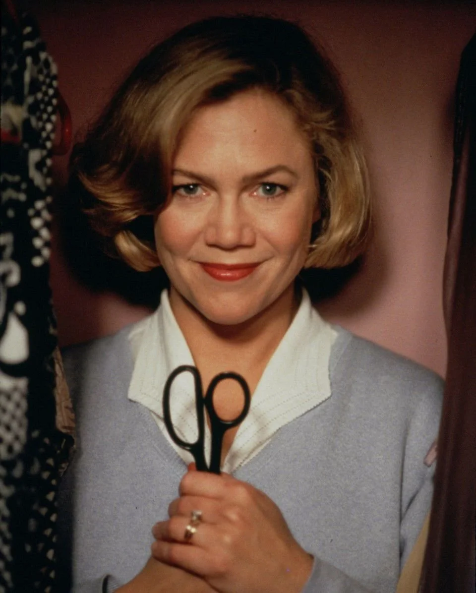 Woman with blonde, shoulder-length hair holding a pair of scissors, smiling, wearing a light gray sweater over a white collared shirt.