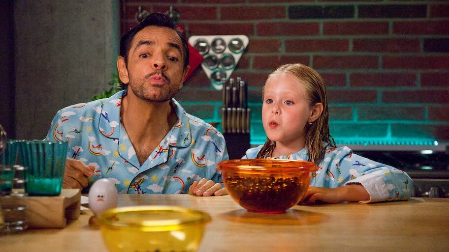A man and a young girl sitting at a kitchen counter with a bowl of cereal in front of them, both making surprised or curious expressions.