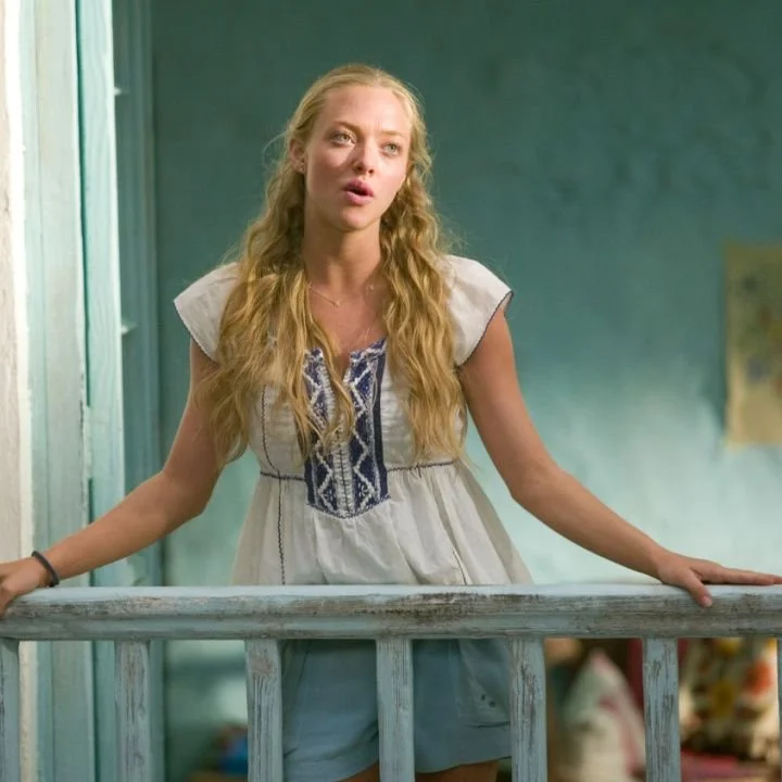 A young woman with long blonde wavy hair standing on a balcony, wearing a white dress with embroidered details, looking thoughtfully to the side.
