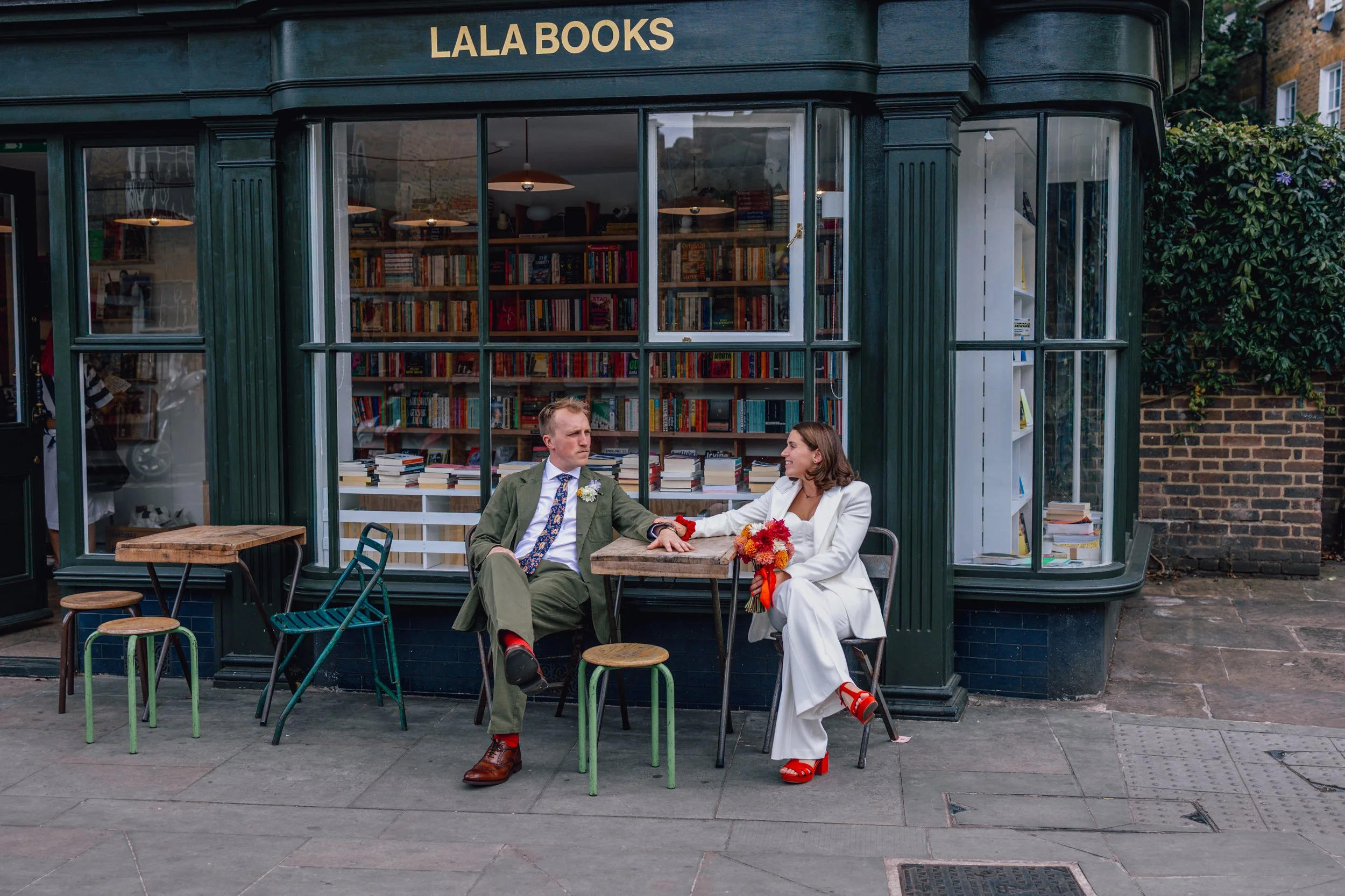 A woman in a white suit and red heels and a man in a green suit with red socks sit outside LALA Books, holding hands. The woman is holding a bouquet of flowers and both are looking at each other, suggesting a romantic or wedding scene.