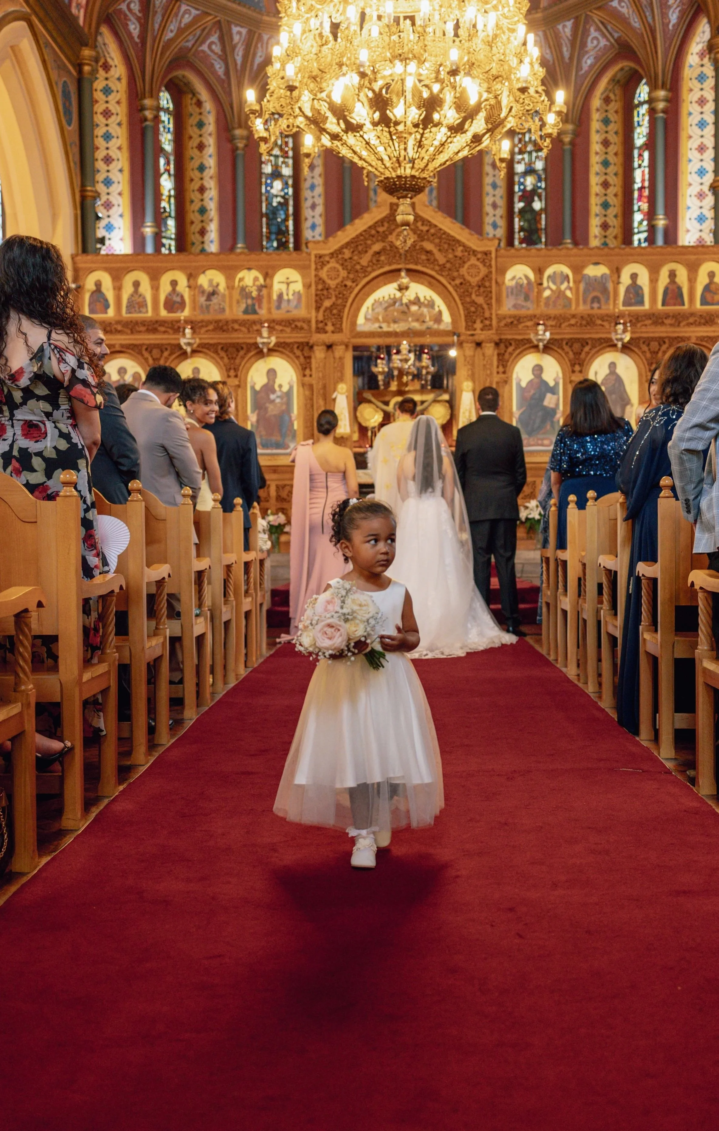 flower girl walking down the aisle towards the camera while a couple get married behind her