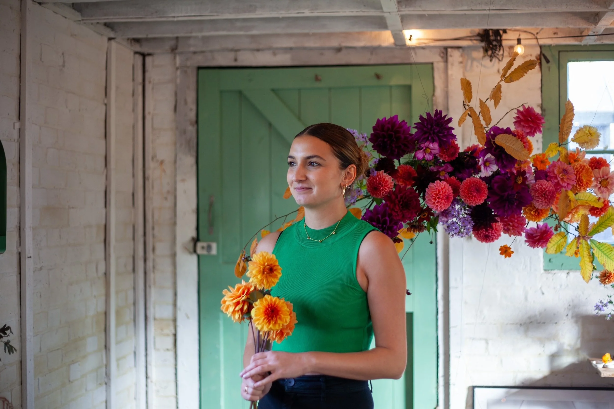 A woman in a green sleeveless top holding a small bouquet of orange and yellow flowers, standing next to a large colorful flower arrangement in an indoor space with brick walls and green doors.