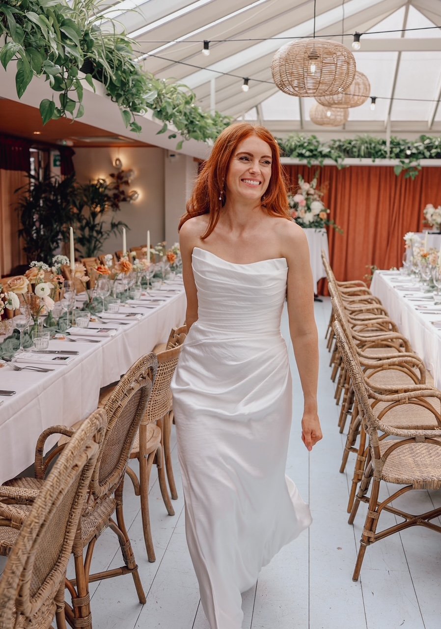 A smiling woman with red hair wearing a white strapless dress walking through a decorated indoor space with a long dining table, floral arrangements, and wicker chairs.