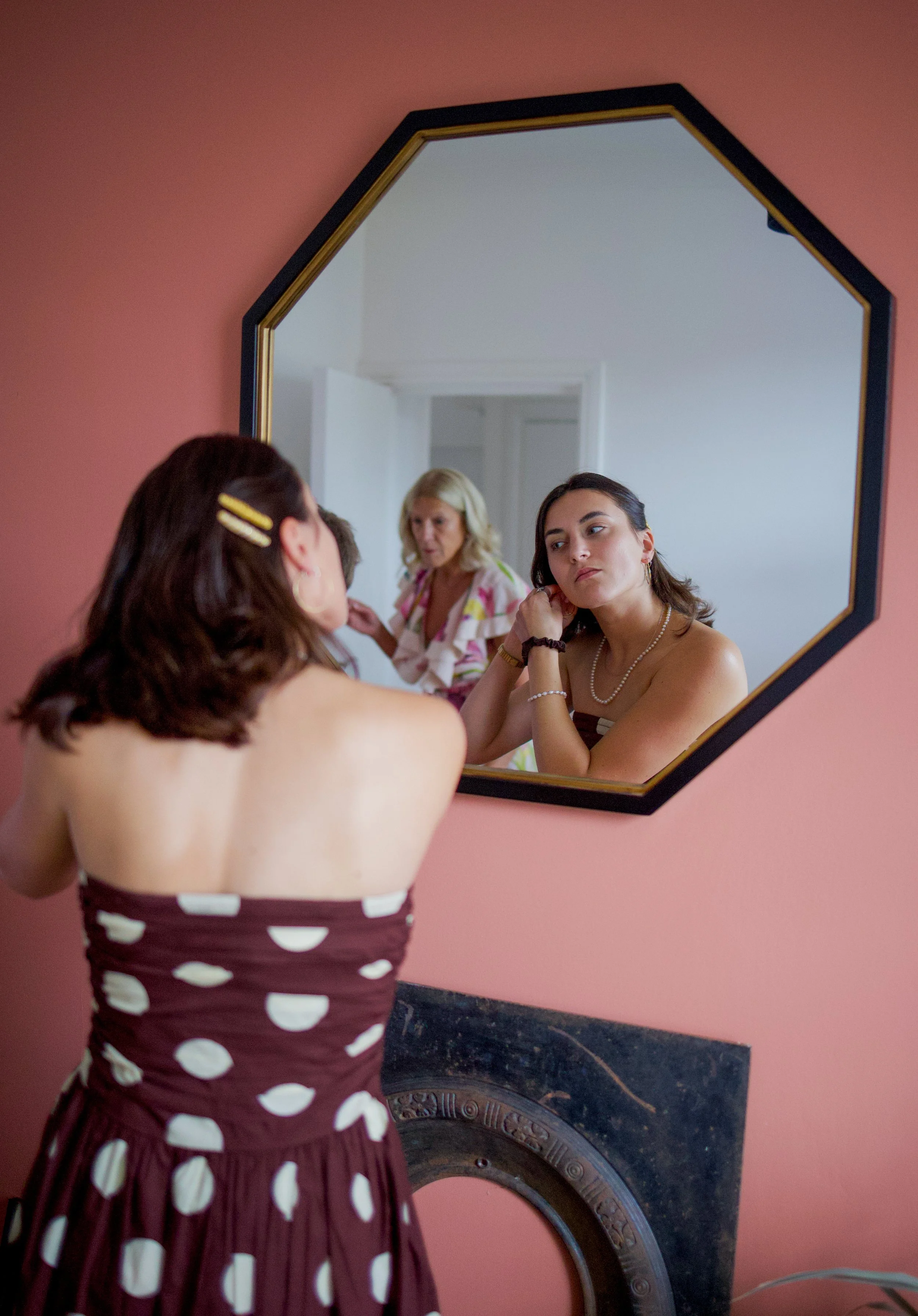 A woman with dark hair and hair clips looks at her reflection in a mirror as another woman, with blonde hair, watches her from behind.