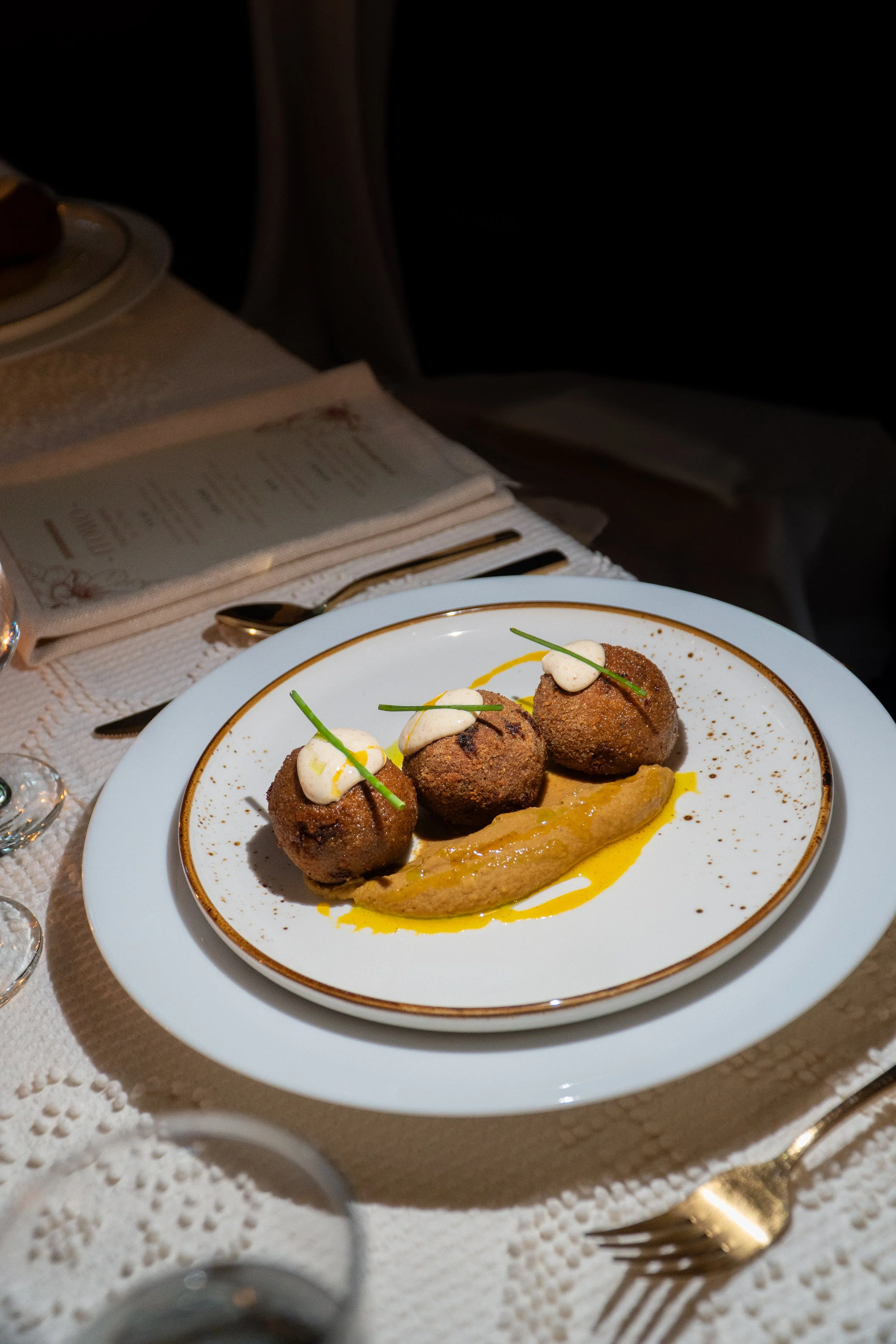 Plate with three arancini balls garnished with herbs, served with a creamy sauce and drizzled with olive oil, on a white plate with a gold rim, placed on a table with a menu, cutlery, and glasses in the background.