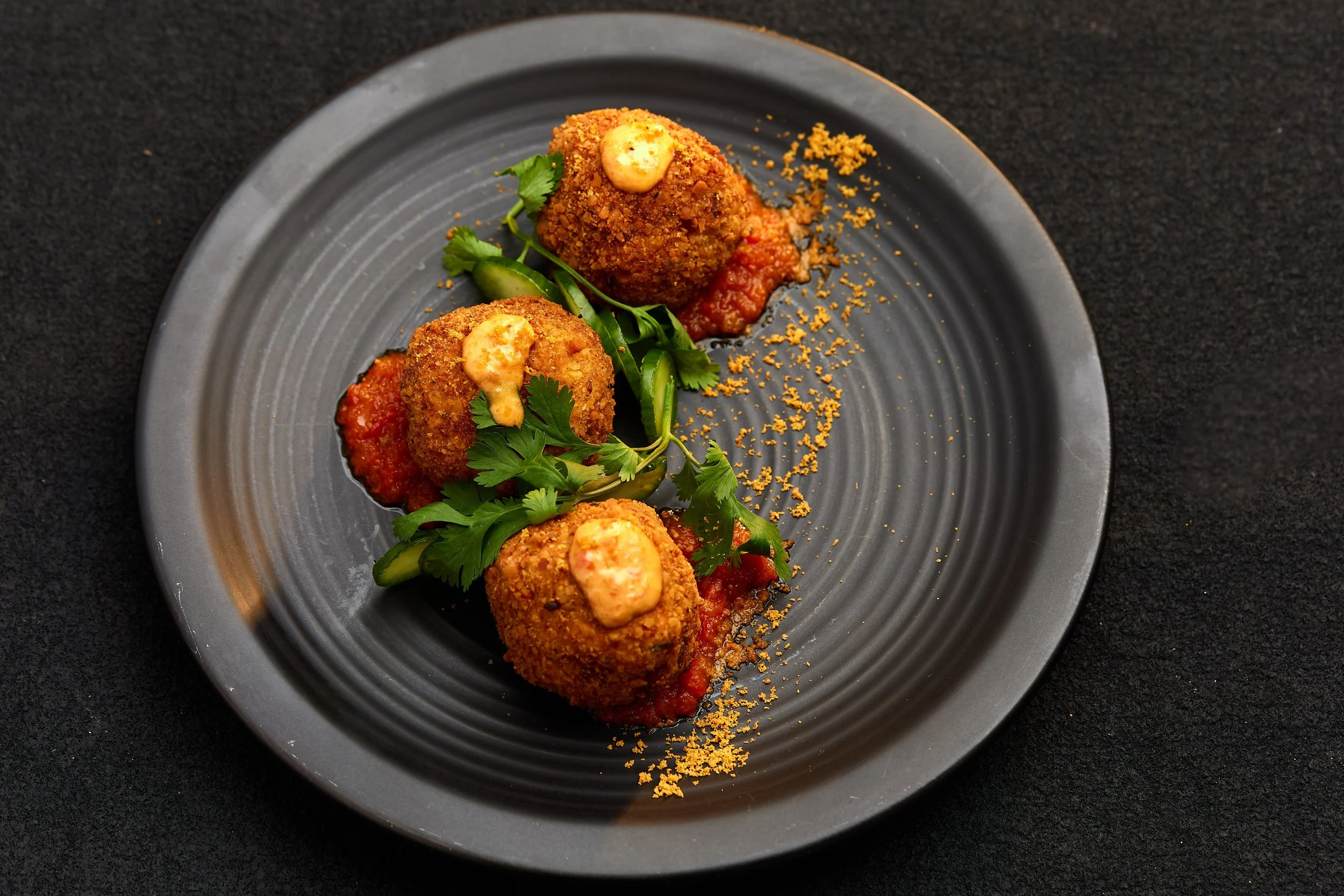 Three fried croquettes topped with sauce on a black plate, garnished with cilantro and green herbs, served with tomato sauce.