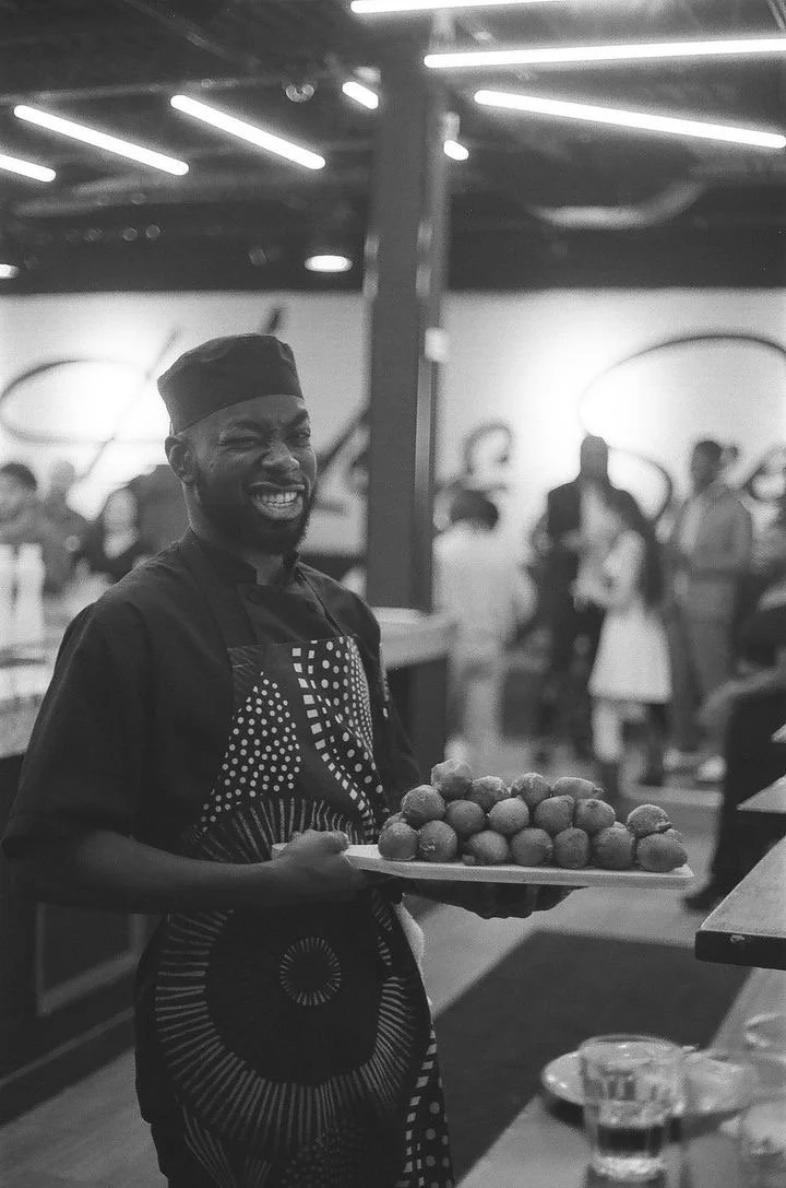 A chef, smiling and holding a tray of food in a busy restaurant or event space.