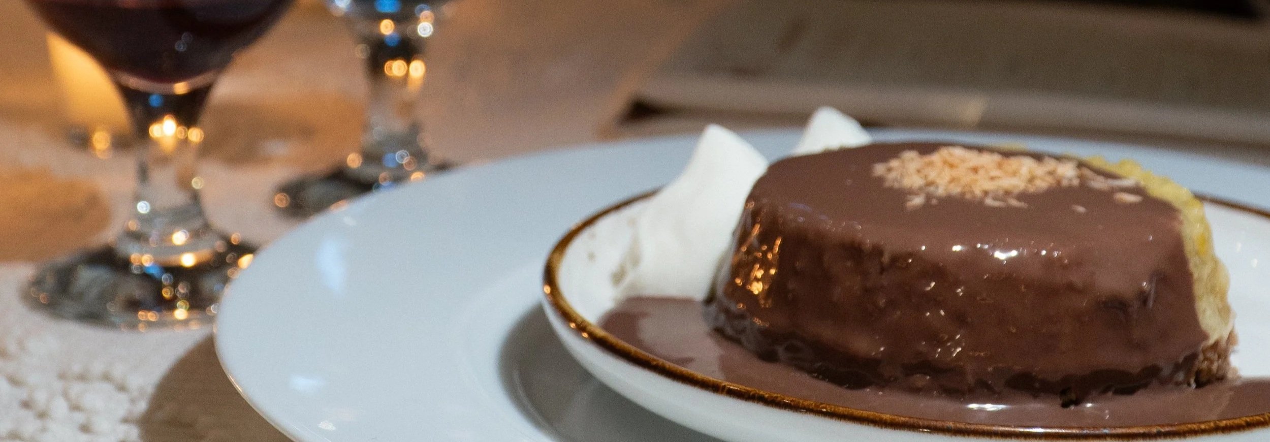 Chocolate cake on a white plate with whipped cream and a glass of red wine in the background.