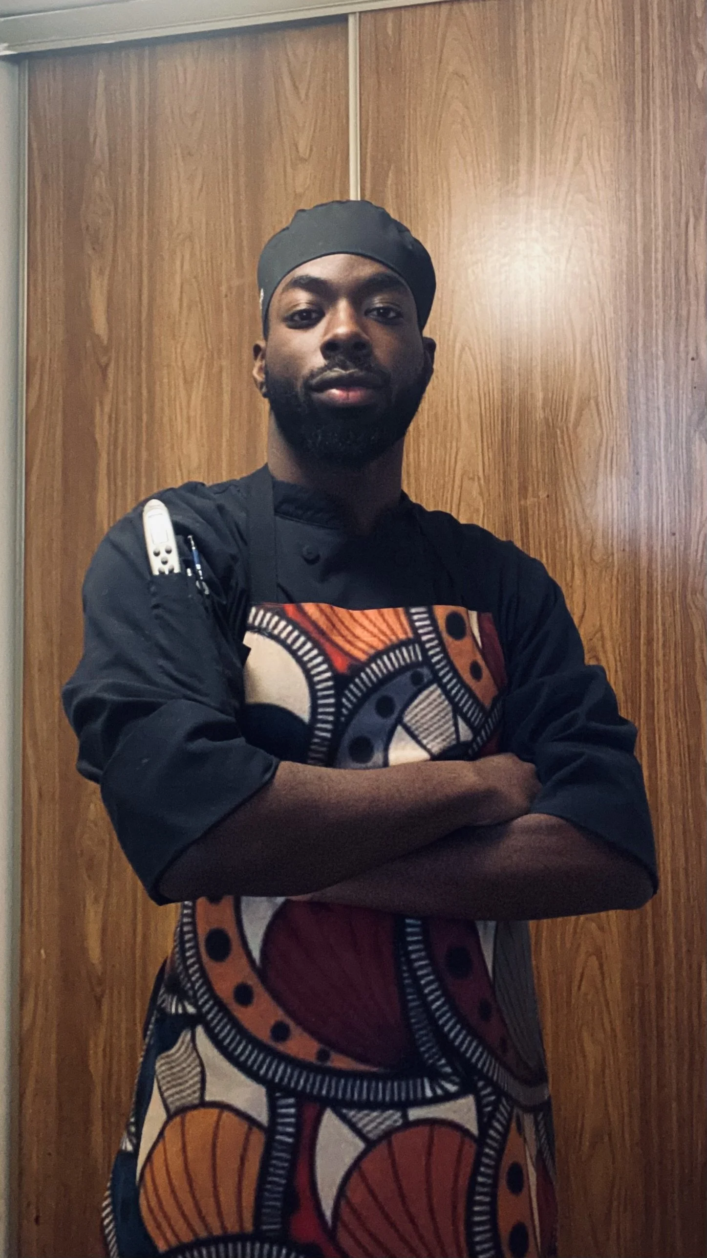 A male chef wearing a black chef's uniform with colorful African print apron, black head covering, standing with arms crossed against a wooden wall.