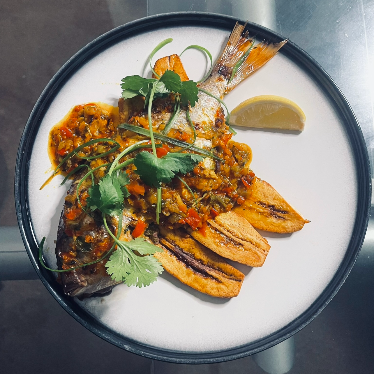 Grilled fish topped with vegetable sauce, garnished with cilantro, served with grilled plantains, lemon wedge, and roasted sweet potato on a white plate