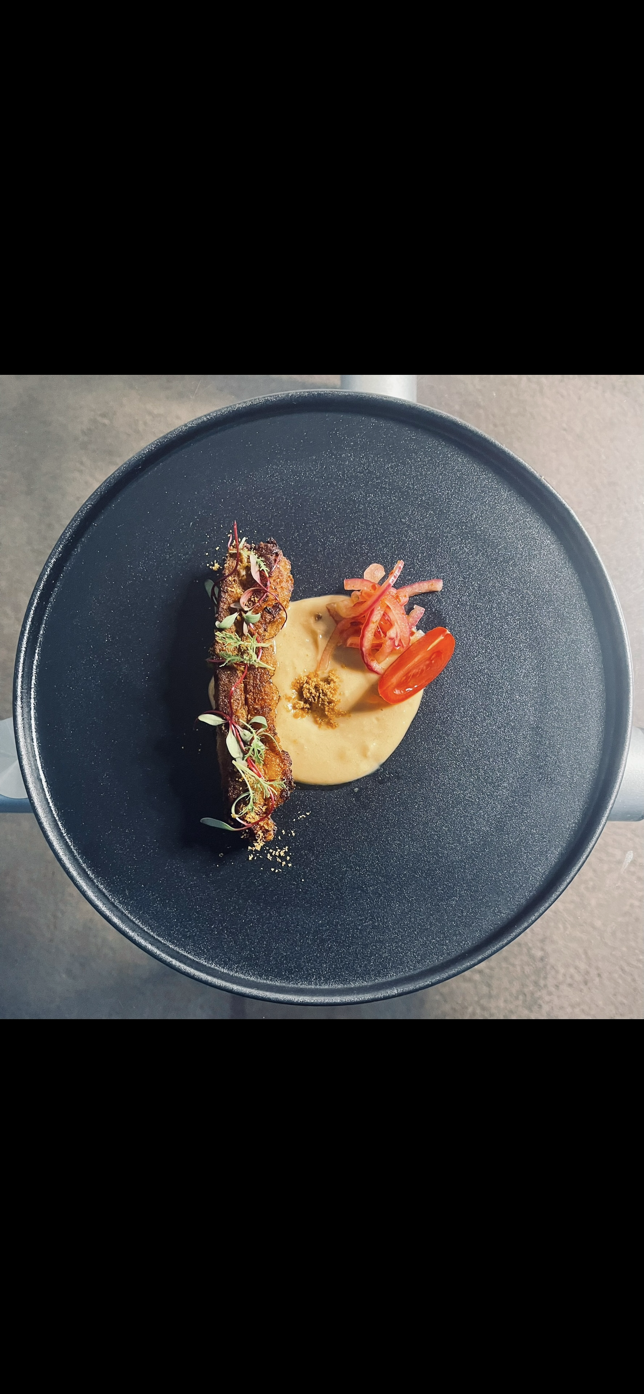 A plated gourmet dish featuring a crispy meat or fish fillet garnished with microgreens, served with a smooth beige sauce, cherry tomato, pickled red onions, and some finely chopped herbs on a dark, round plate.