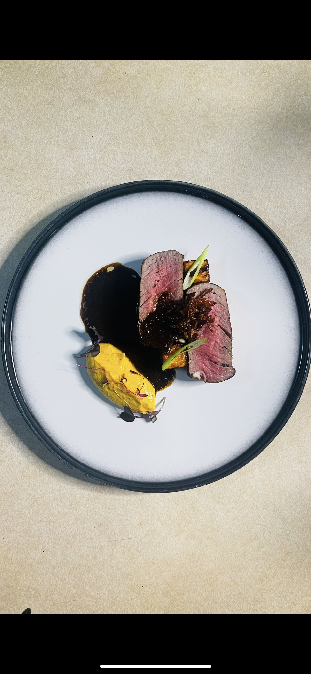 A plated gourmet meal featuring two slices of medium-rare steak with a dark sauce, garnished with green herbs, served with a dollop of bright yellow sauce or puree, on a round black and white plate.
