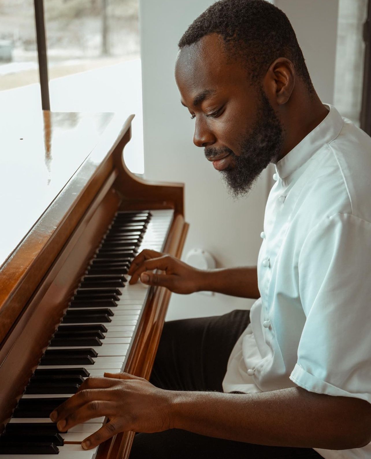 A man with a beard, wearing a white chef coat, playing a wooden upright piano indoors near a window.