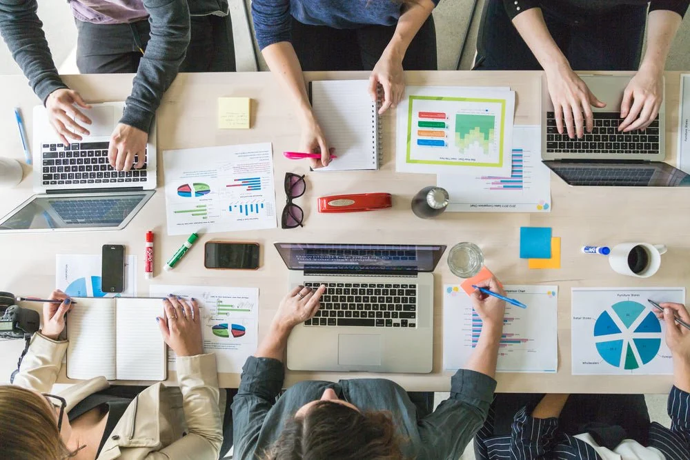 Top-down view of a business meeting with six people working on laptops, charts, and documents at a conference table.