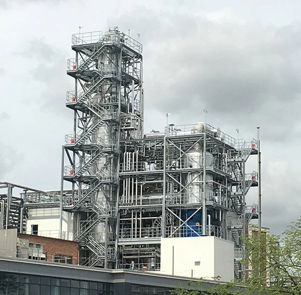 Industrial chemical processing plant with metal structures, pipes, and staircases under cloudy sky.
