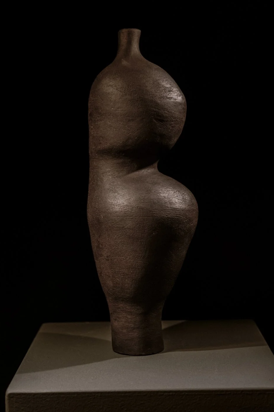 Eine dunkle, formschöne Skulptur eines menschlichen Körpers, die vage Frauenfigur darstellt, auf einem weißen Sockel gegen schwarzen Hintergrund.