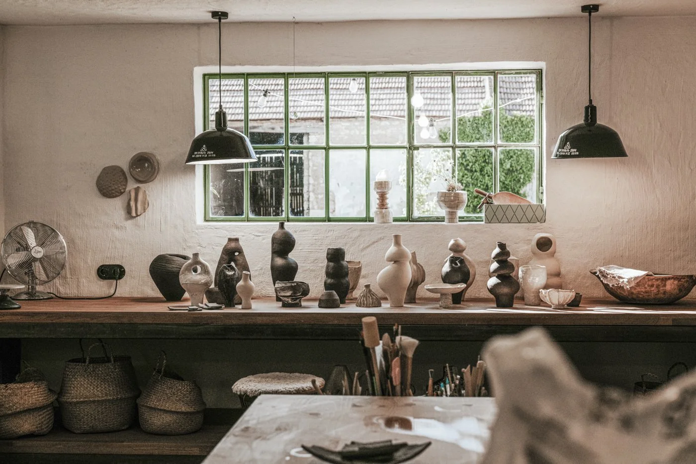Blick in eine Keramikwerkstatt mit einer Werkbank, auf der verschiedene Keramikwaren an der Wand und auf der Fensterbank stehen. Über dem Fenster hängen zwei schwarze Lampen.