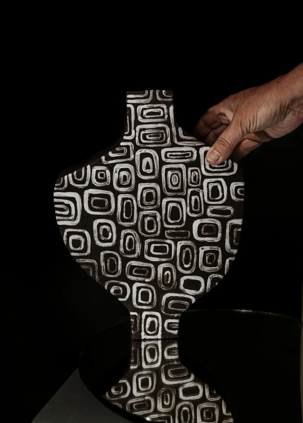Eine Hand hält eine schwarze Vase mit weißen geometrischen Mustern, umgeben von einem schwarzen Hintergrund.