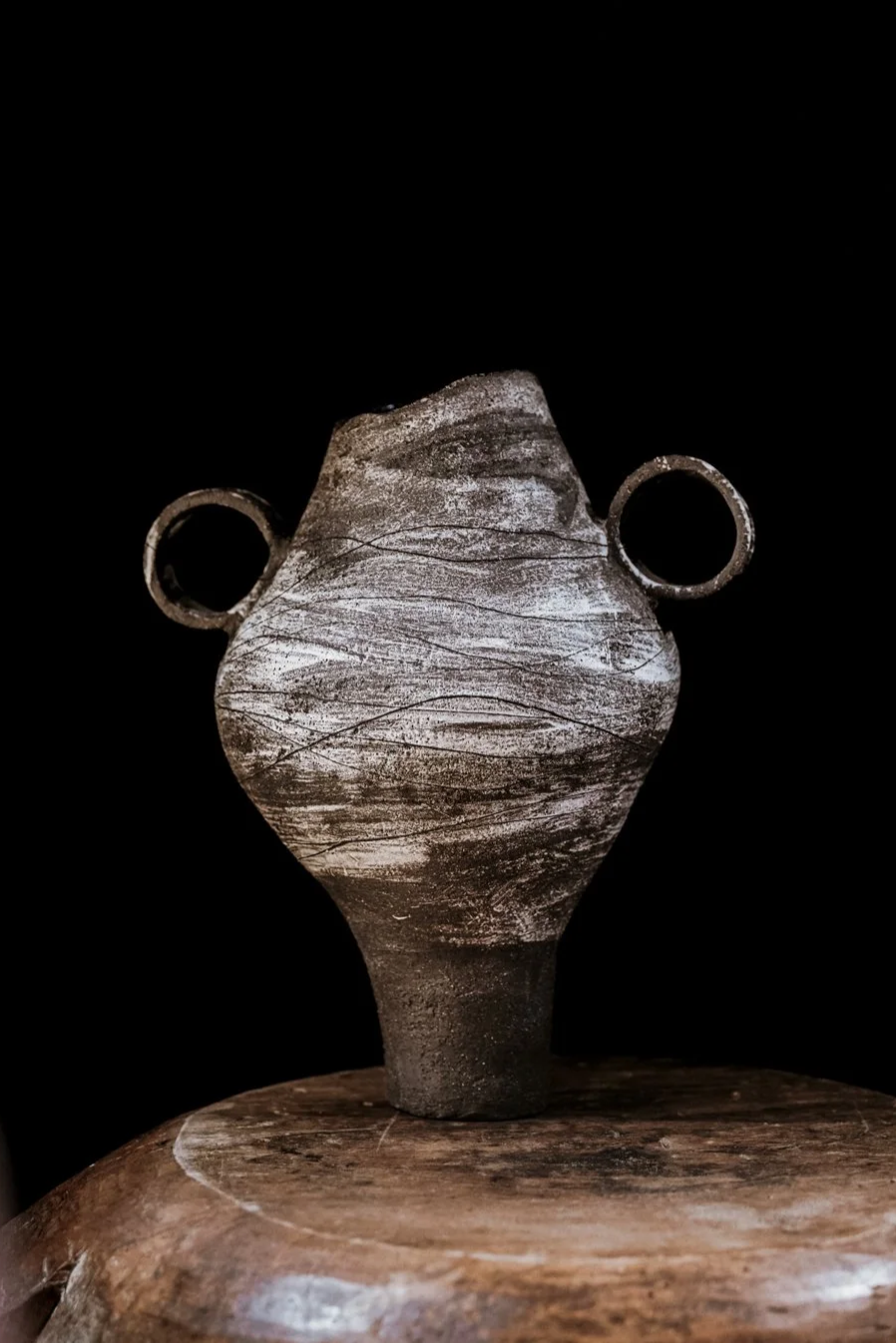 Antike Keramikvase mit zwei seitlichen Henkel auf einem Holztisch vor schwarzem Hintergrund.