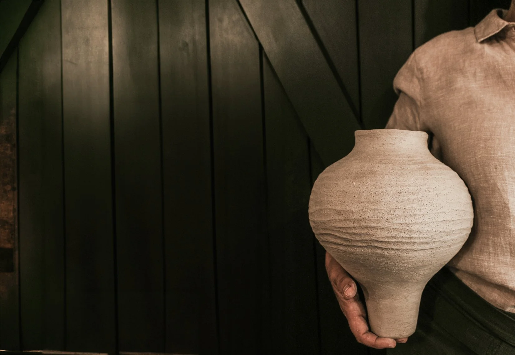 Person hält eine große, ungeglättete Tonvase vor einer schwarzen Wand.