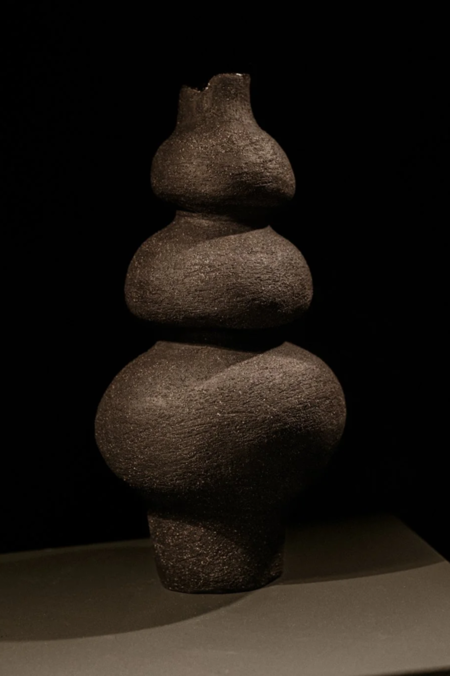 Eine schwarze, abstrakte Skulptur aus grobem Material, die aus vier unterschiedlich großen, kugelartigen Formen besteht, die übereinander gestapelt sind.