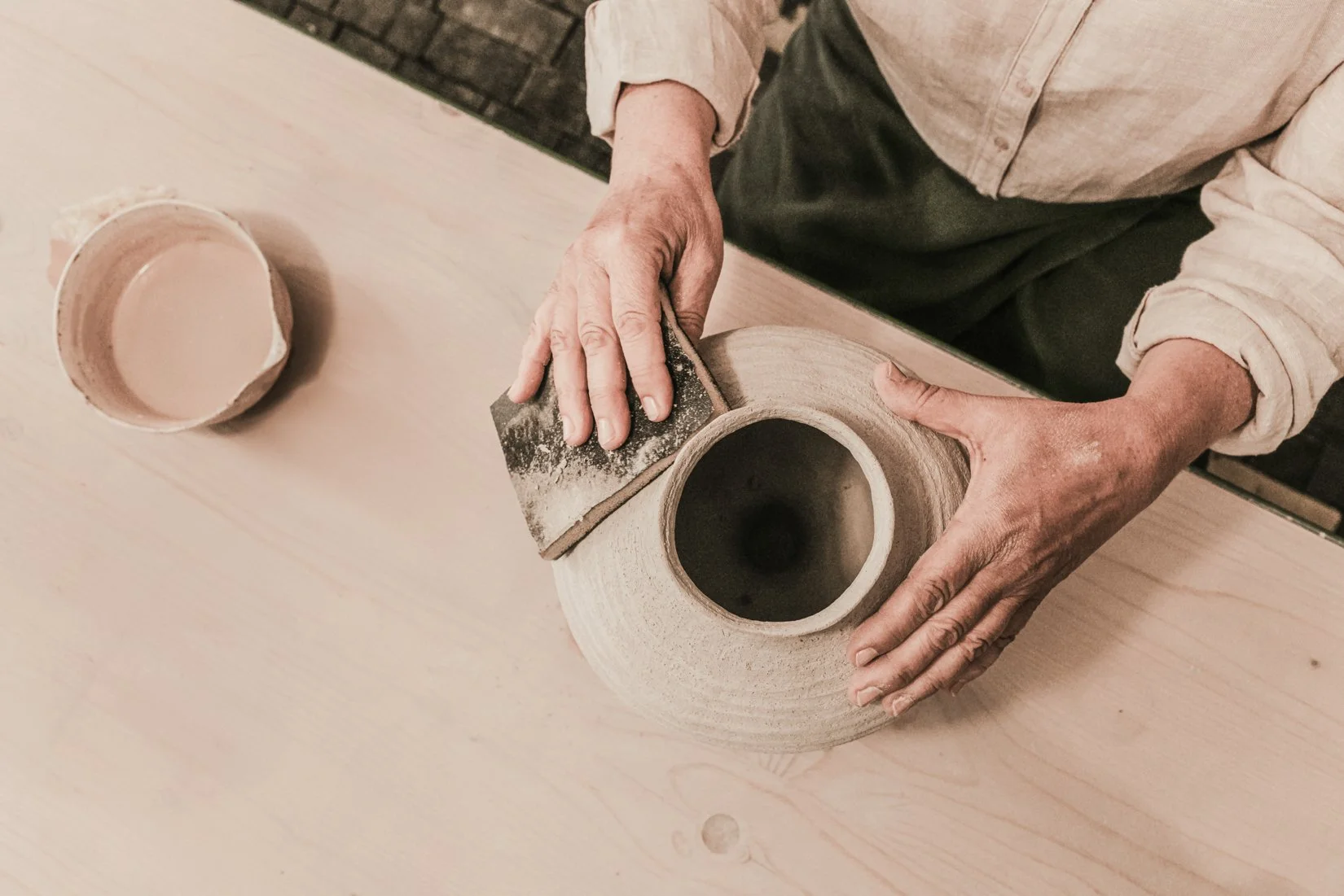 Person schleift die Oberfläche eines Keramiktopfes mit Schleifpapier auf einem Tisch.