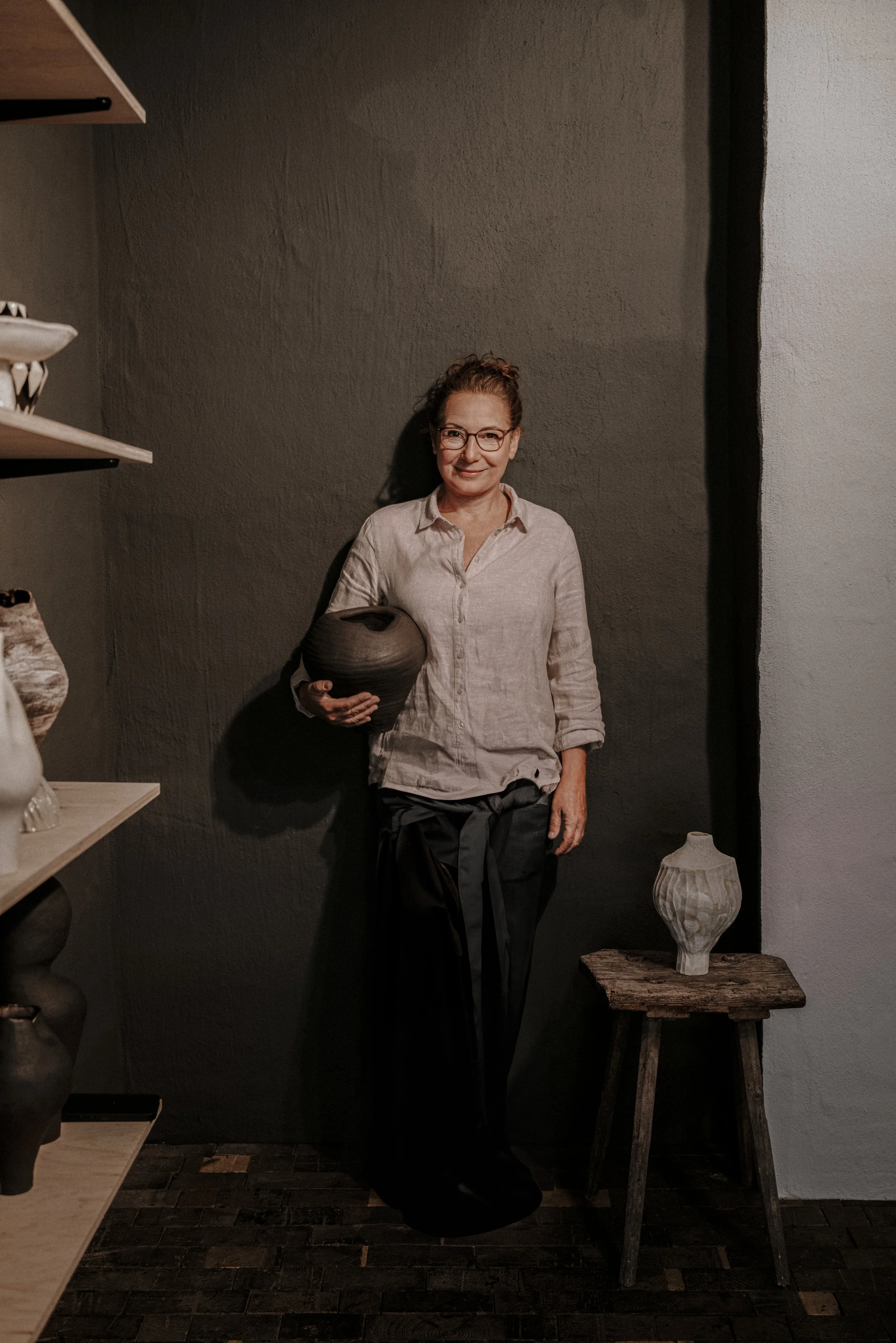 Eine Frau mit Brille hält einen schwarzen Keramiktopf in ihrer linken Hand und steht vor einer dunklen Wand, umgeben von Keramikvasen auf Regalen und einem Holztisch.
