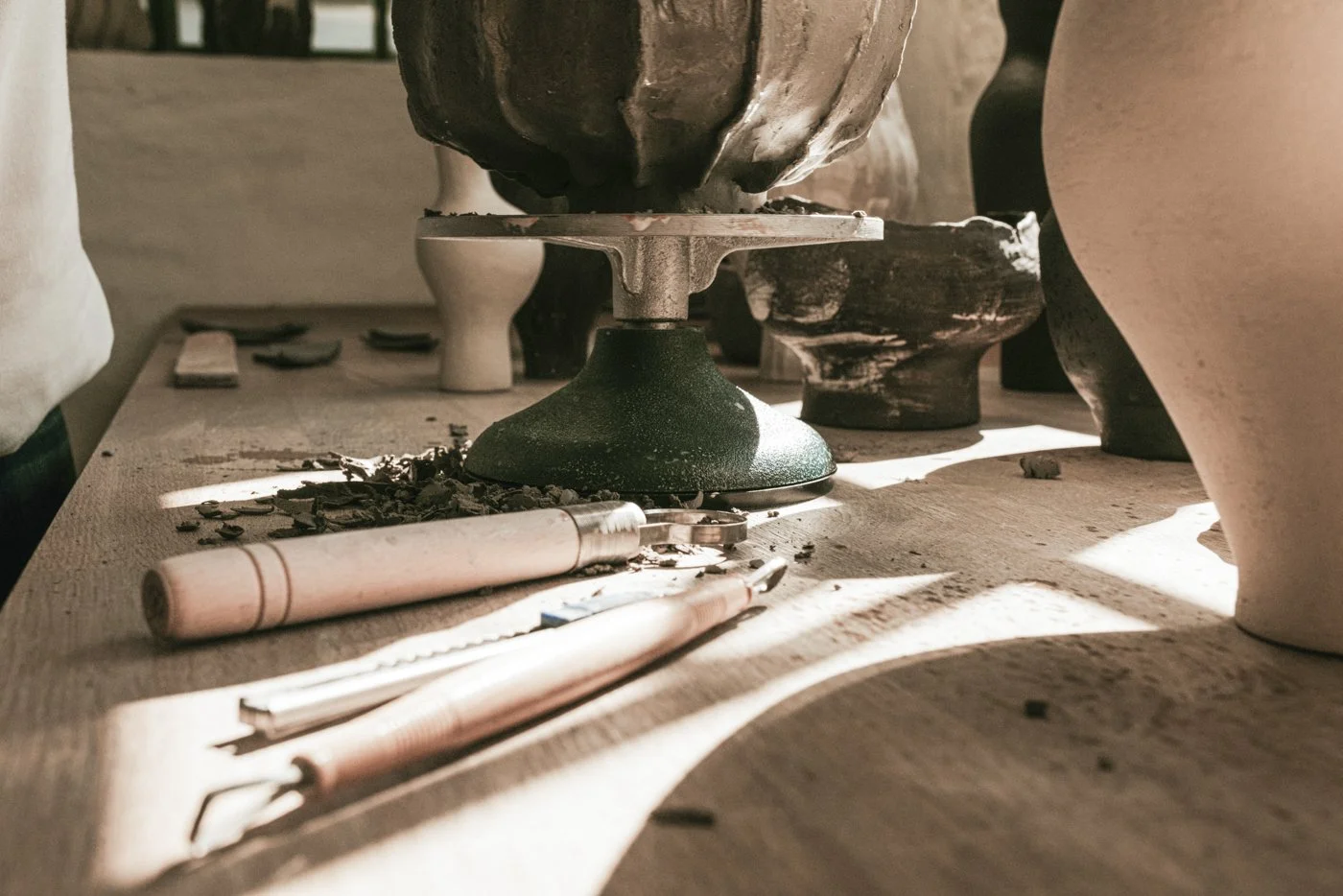 Töpferscheibe im Keramikatelier mit Ton, Trocknungsformen und Werkzeug auf einem Holztisch, durch Sonnenlicht beleuchtet.