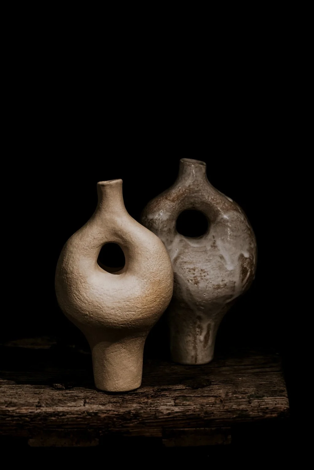 Zwei antike Keramikvasen mit runden Löchern in der Mitte, auf einem alten Holzbrett gegen einen schwarzen Hintergrund.