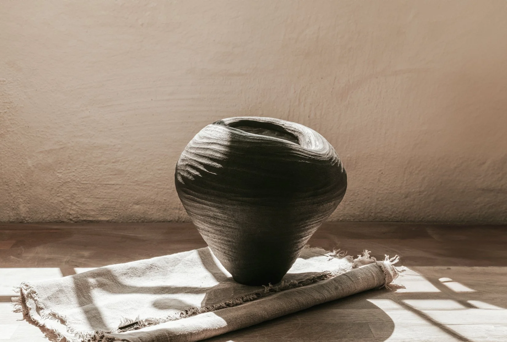 Schwarze, strukturierte Vase auf einer weißen Stoffserviette, im Sonnenlicht gegen eine beige Wand.