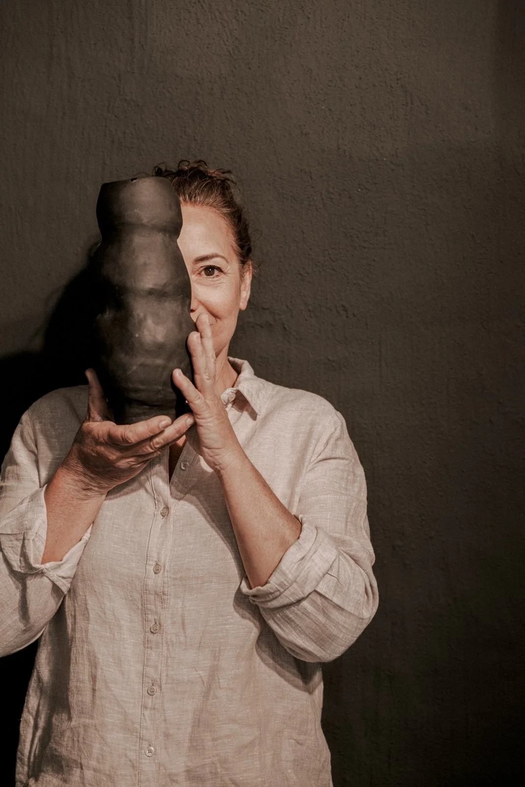 Frau hält eine große, dunkle Tasche vor ihr Gesicht, zeigt nur ein Auge und halb lächelt, vor einer dunklen Wand.