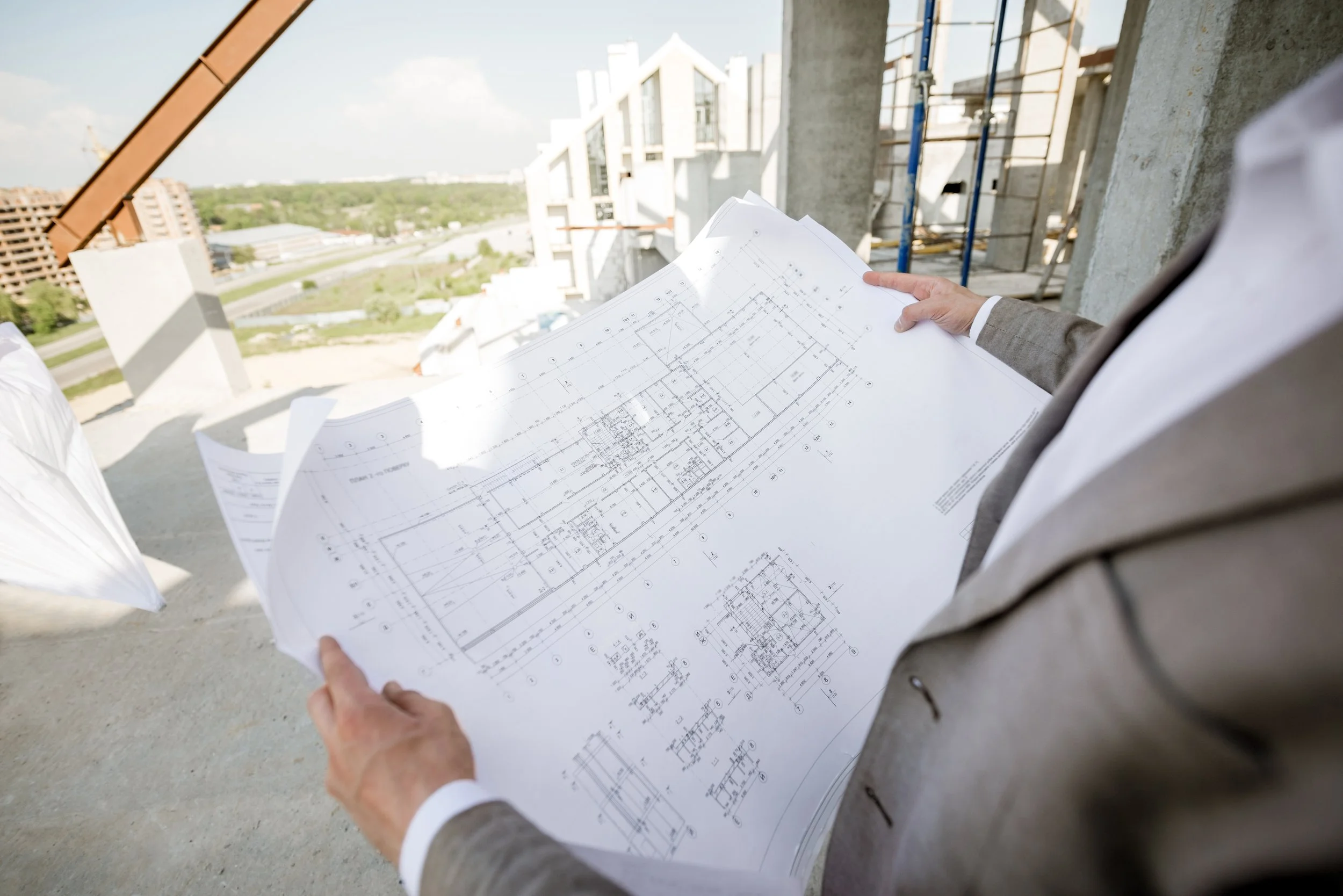 Construction site with a person in a suit holding architectural blueprints, with a building under construction in the background.