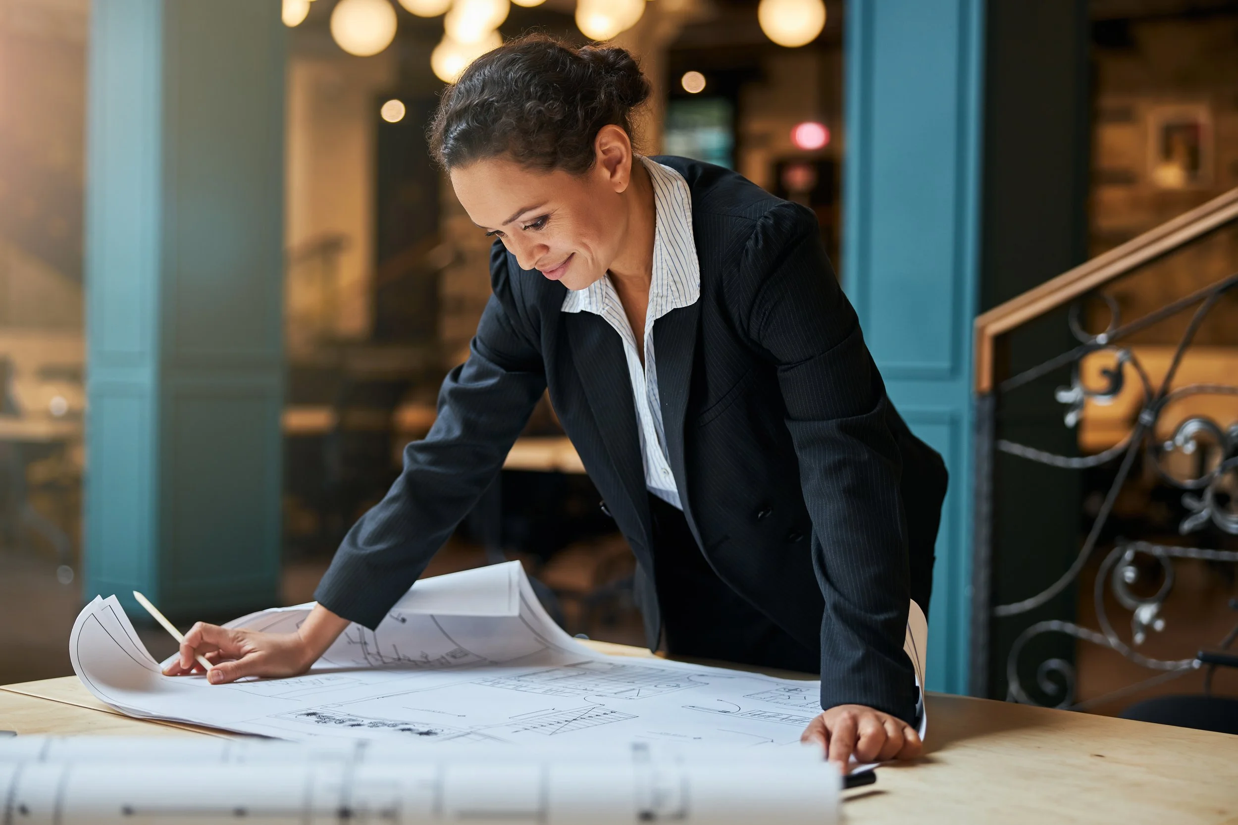 Businesswoman in a dark blazer and striped shirt examining architectural blueprints on a table.